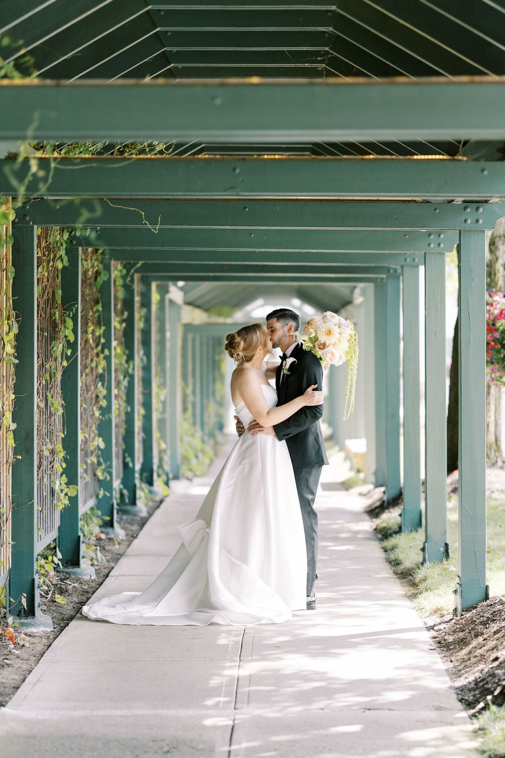 A romantic wedding couple embraces in a long covered walkway with green pillars and natural sunlight streaming in.