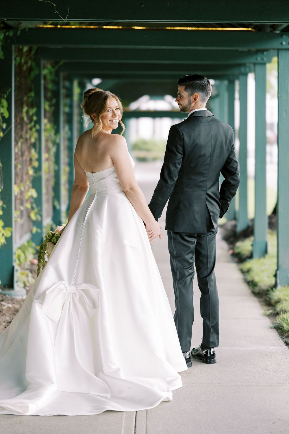Wedding couple walks together under covered outdoor walkway with sunlight filtering through green foliage.