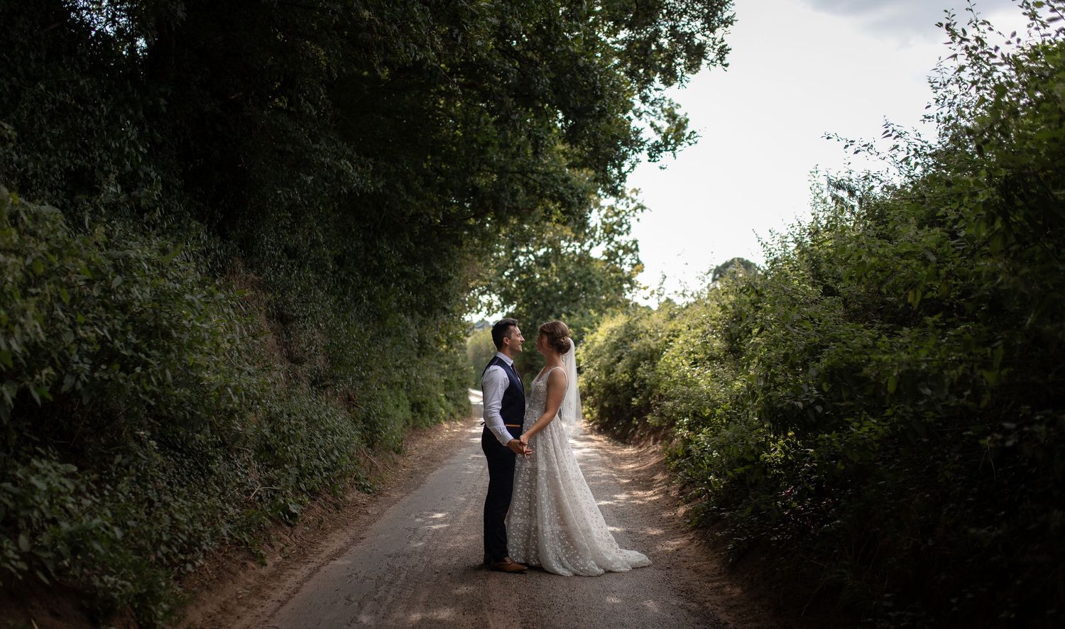 A couple shares an intimate moment together on a dirt path surrounded by lush green trees and foliage.
