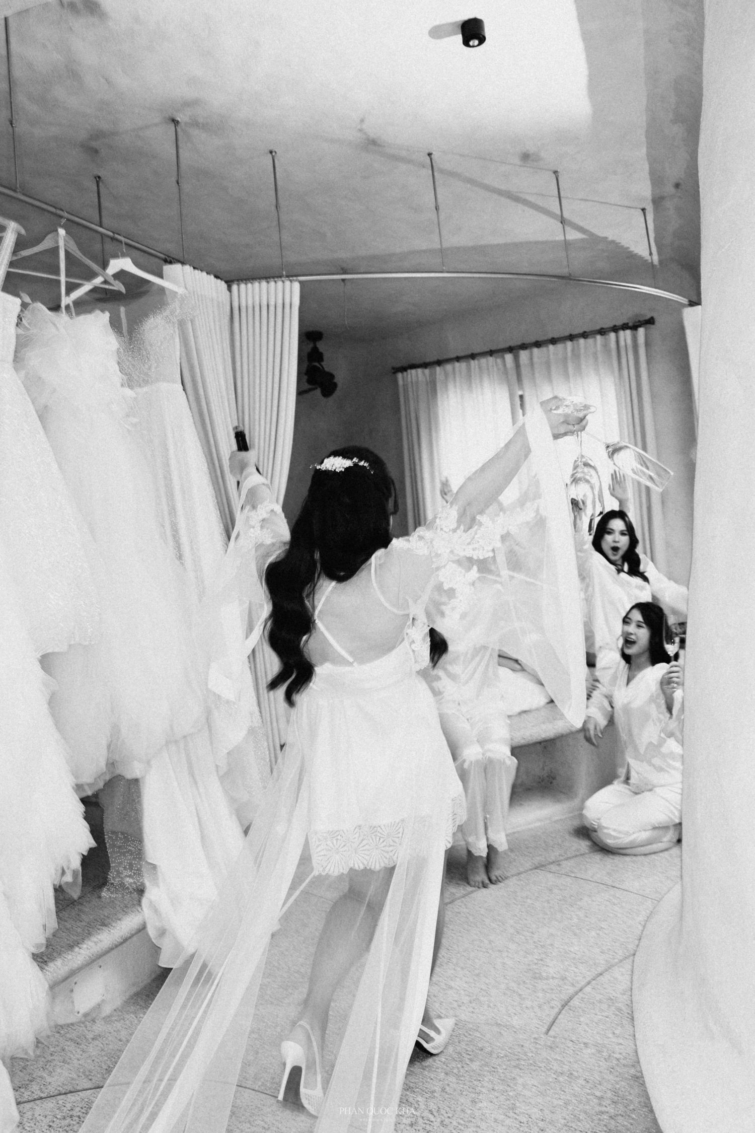 Bride in white lace robe browses wedding dresses in bridal boutique with bridesmaids in black and white photo.