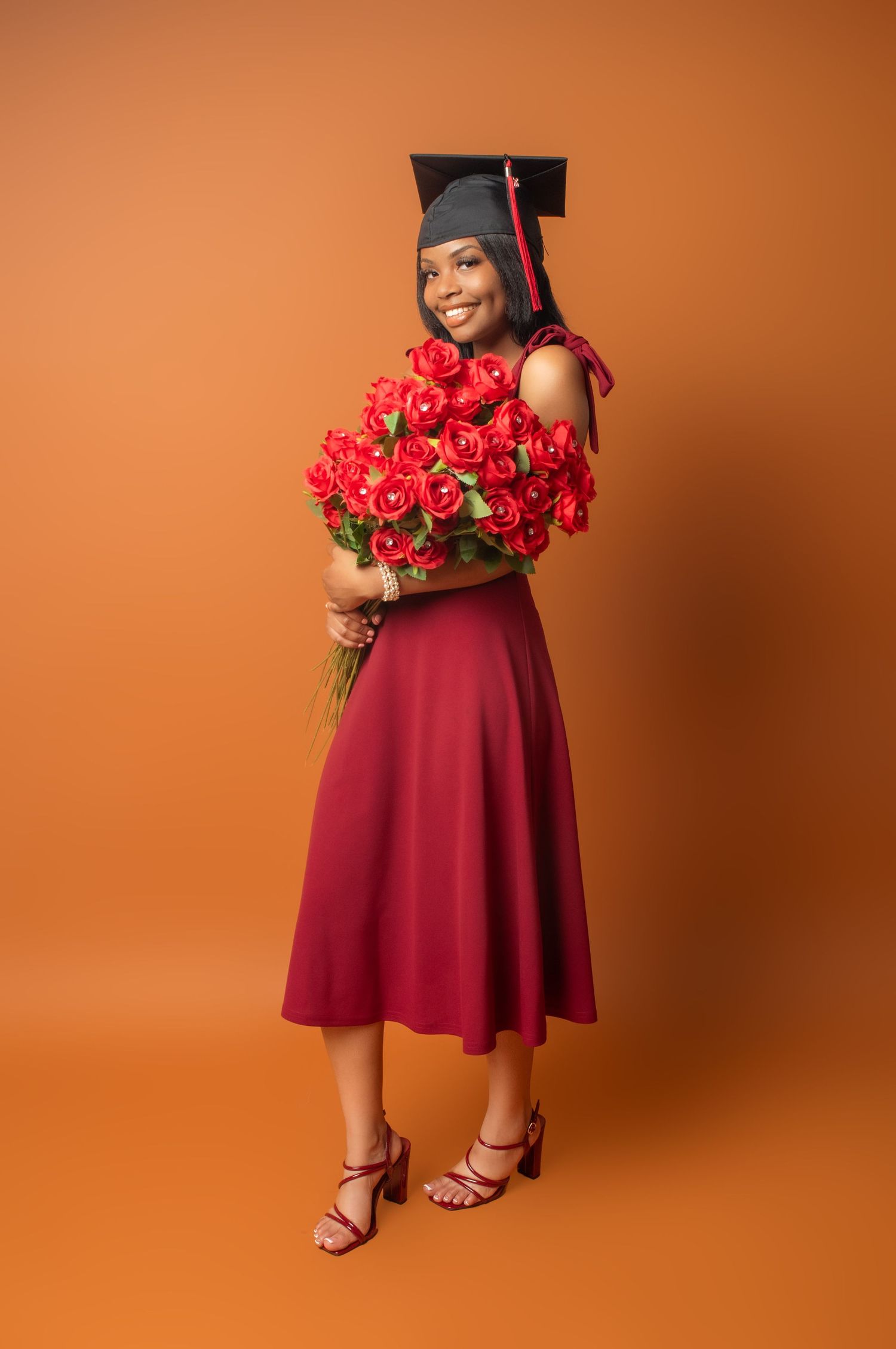 Red roses and burgundy dress create an elegant graduation portrait against an orange backdrop.