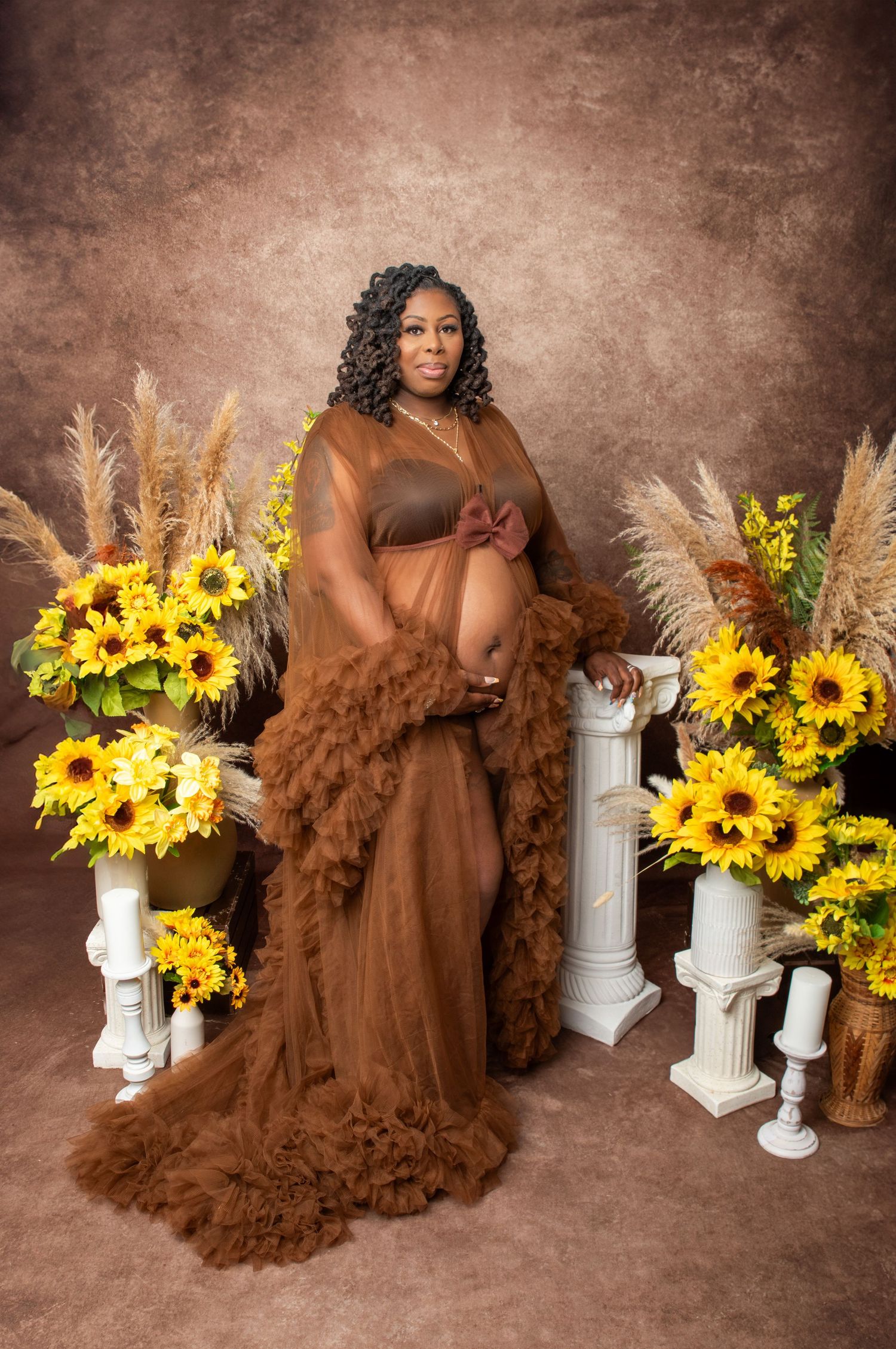 Beautiful maternity photo with brown flowy dress surrounded by sunflowers and pampas grass against sepia backdrop.