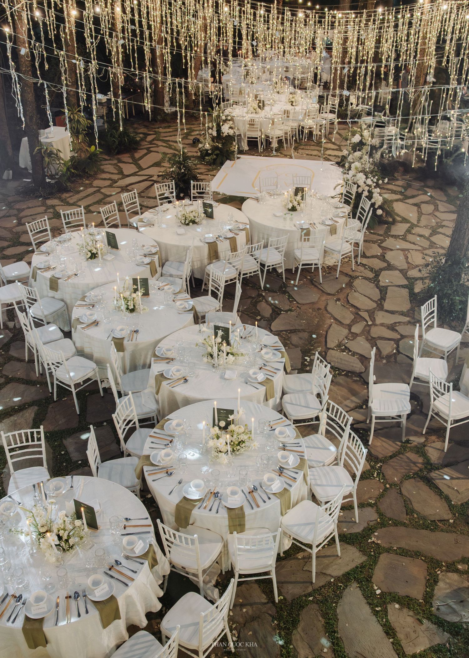 Elegant outdoor wedding reception with round tables, white chiavari chairs, and string lights overhead.