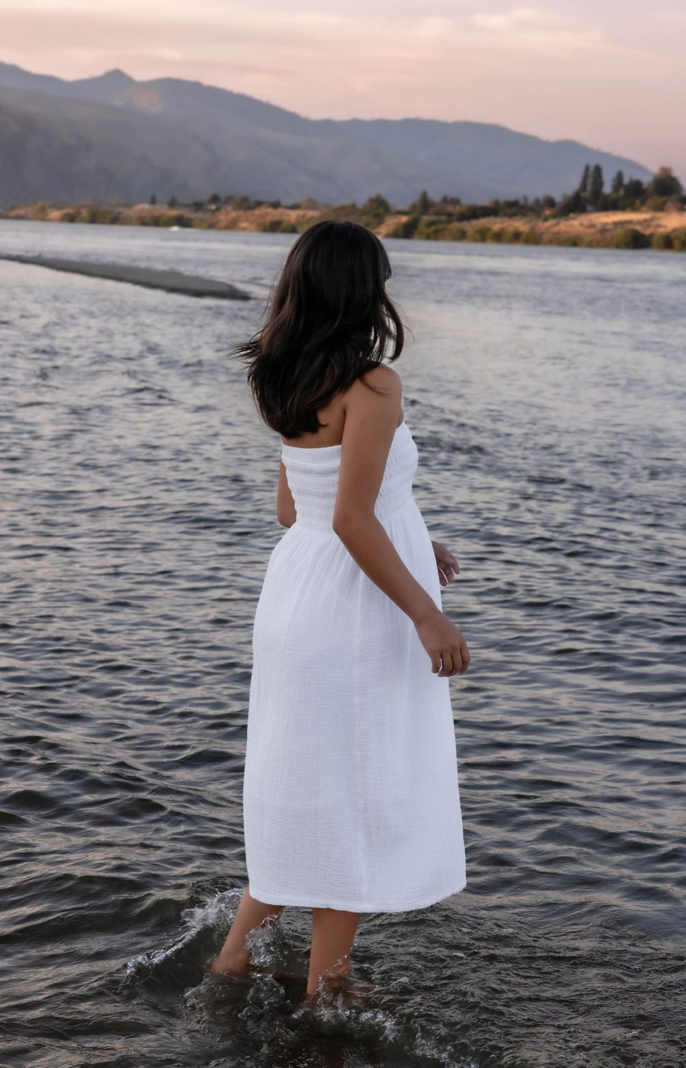 Person in white dress splashing in lake water at sunset with mountains in background.