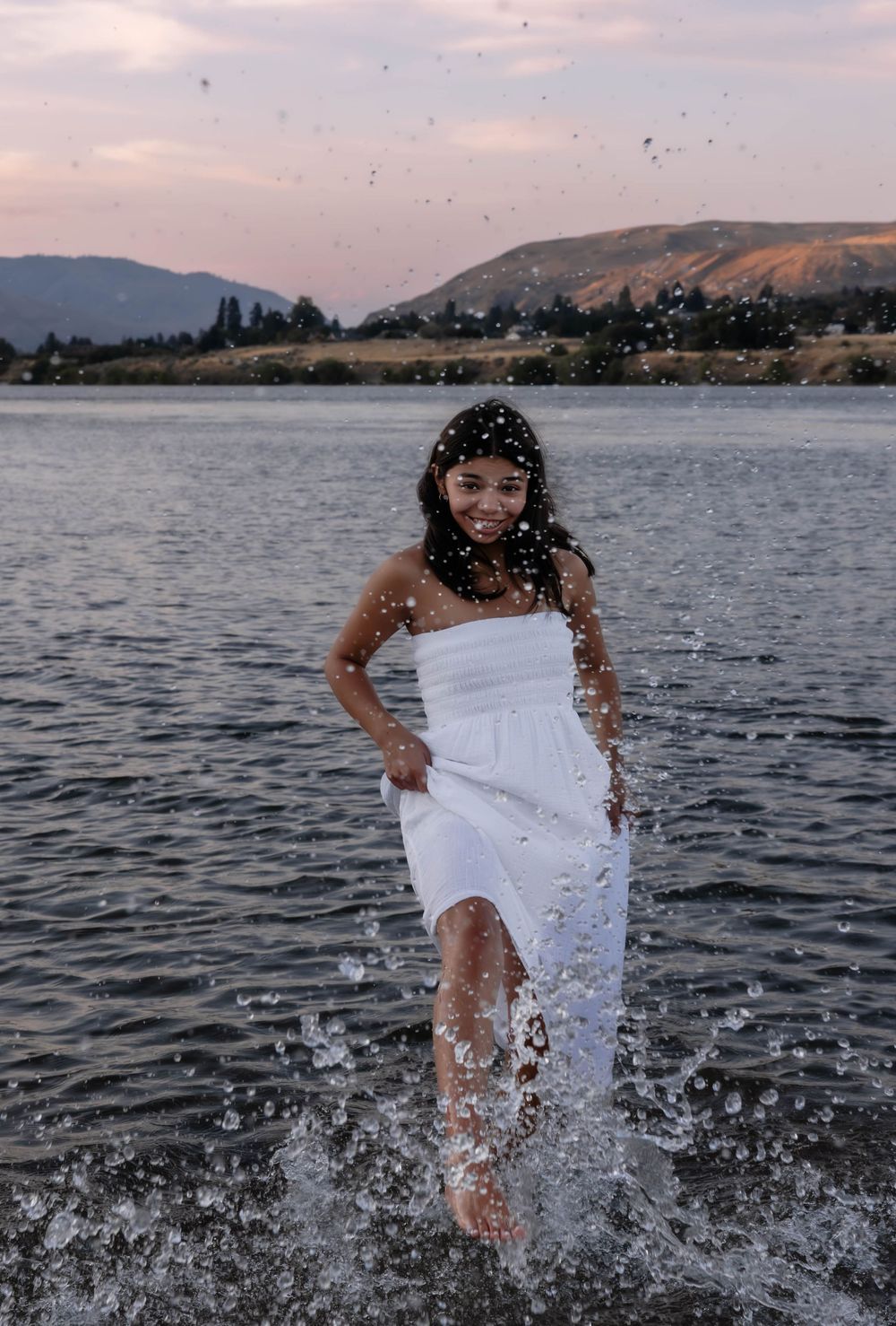 Person playing in lake water wearing white dress during sunset, creating splashes.