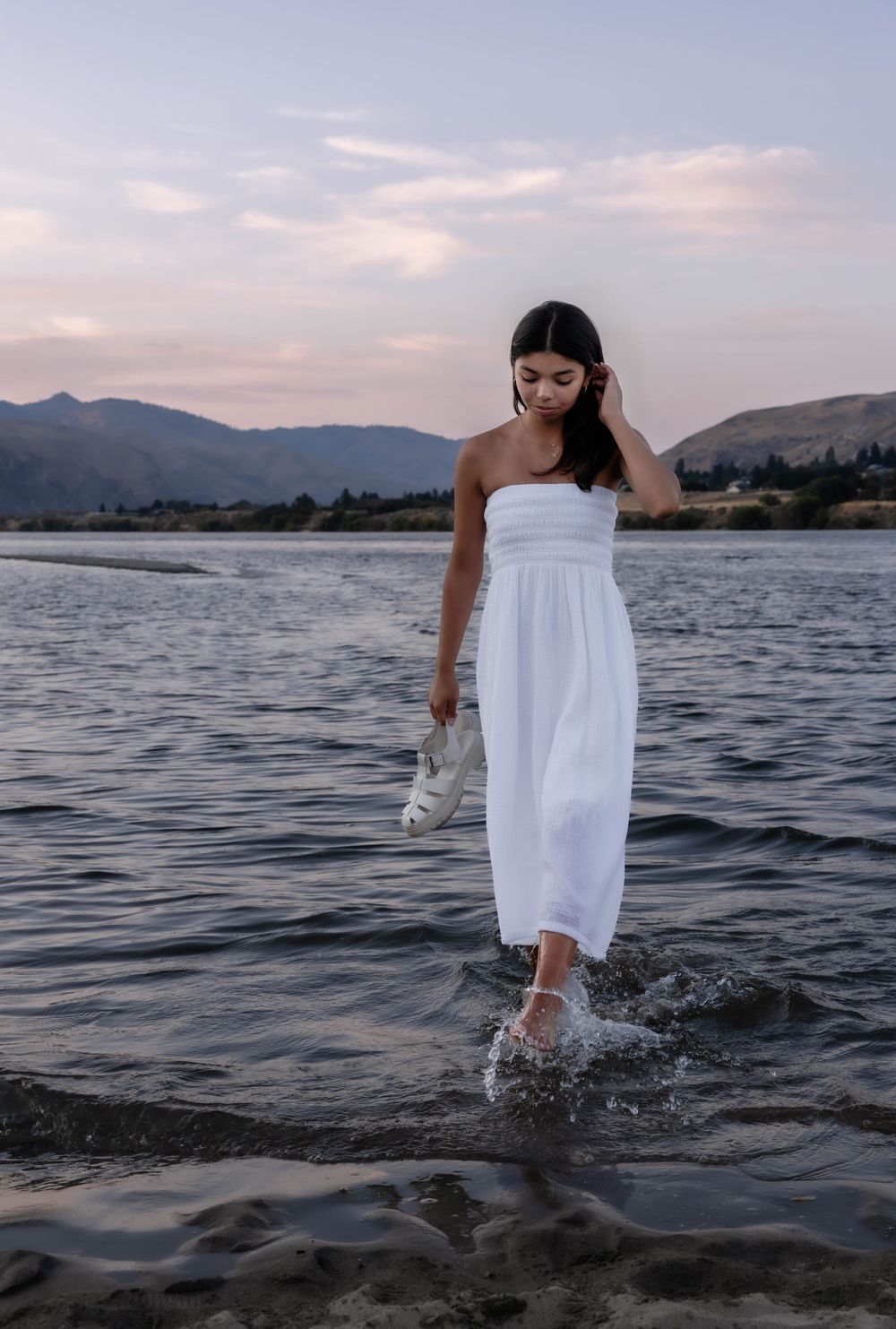 Figure in white dress wading through mountain lake water at dusk.