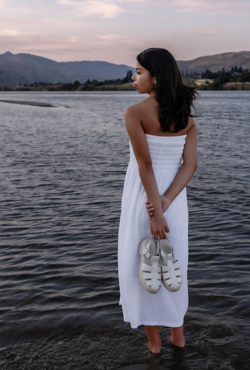 Person in white dress holding sandals while standing in lake water at sunset.