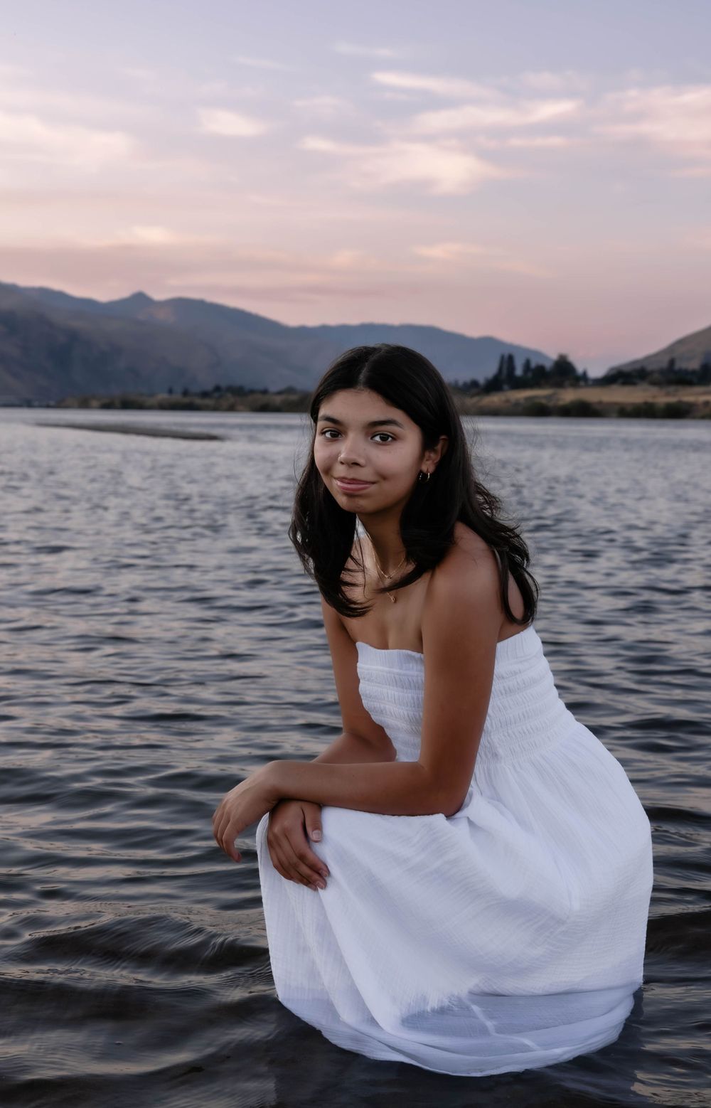 Person in white dress poses by the lake during sunset with mountains in background.