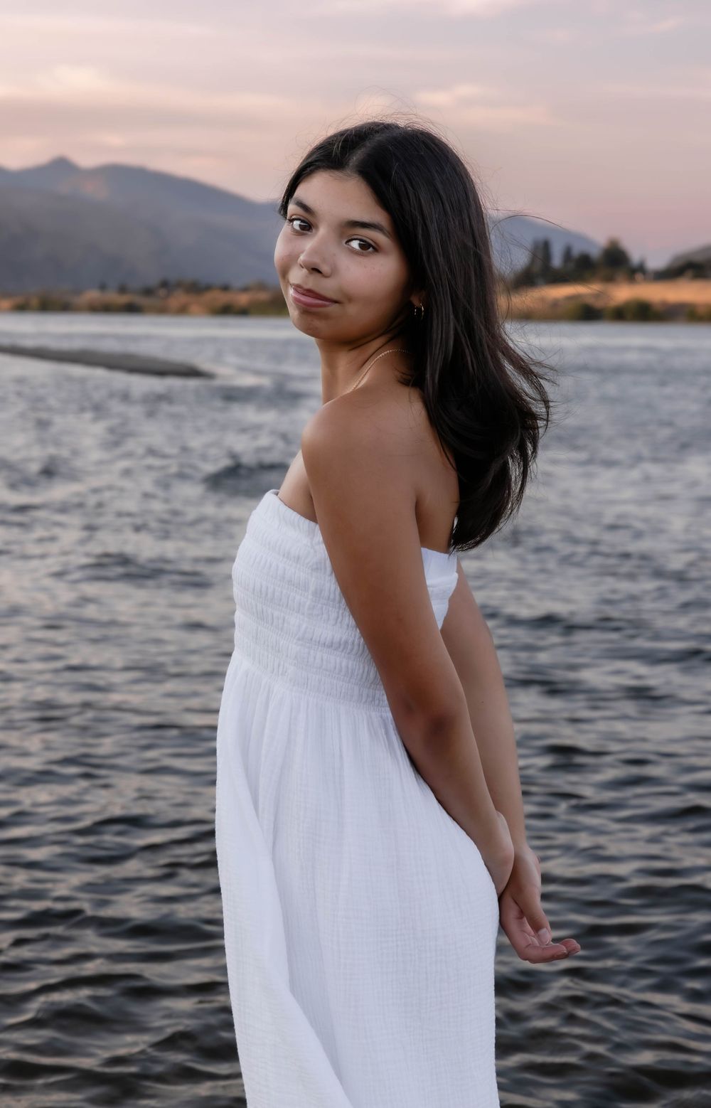 Person in white sundress poses by lake with mountain landscape at dusk.