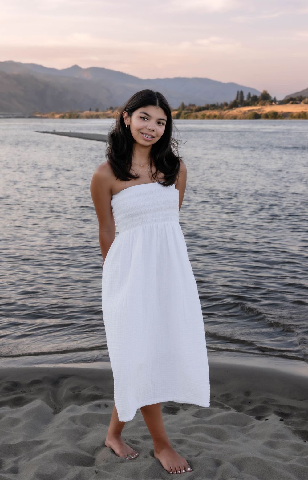 Person in white sundress standing on beach near mountain lake.