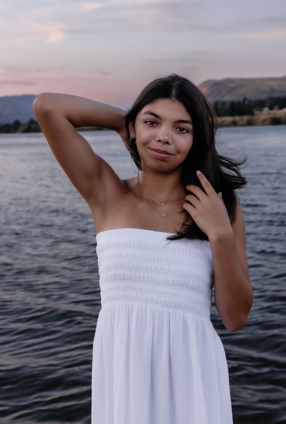 Person in white summer dress poses against sunset lake and mountain backdrop.