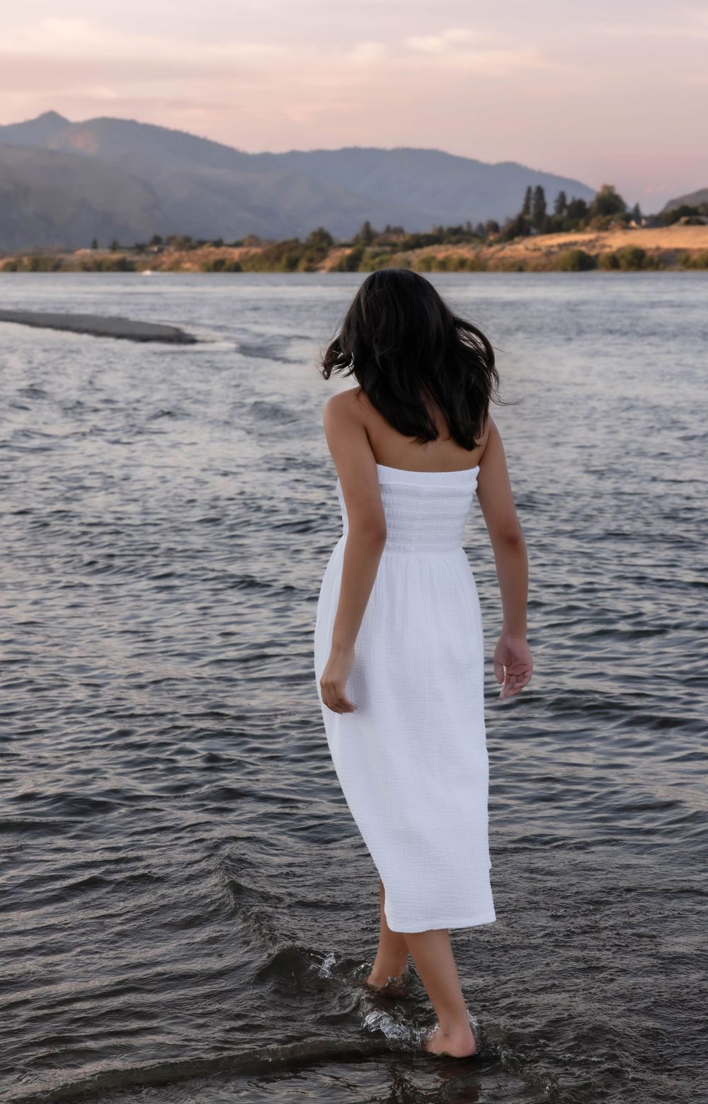 Person in white dress wading into a mountain lake at sunset.