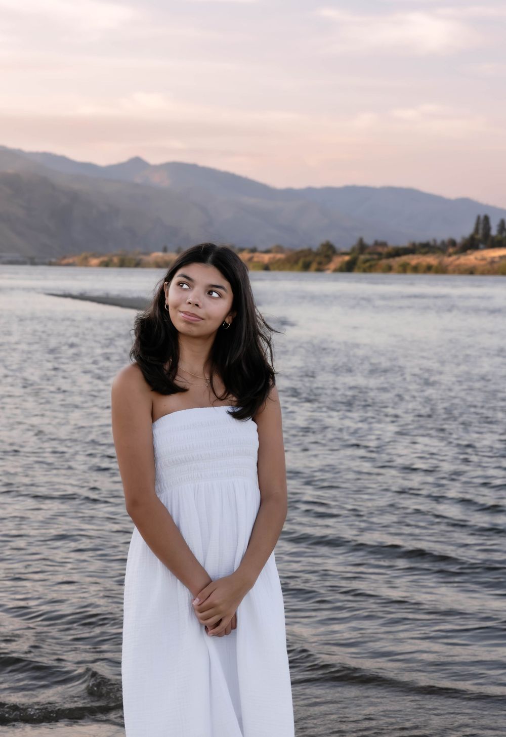 Person in white dress poses by a scenic lake with mountains in background.