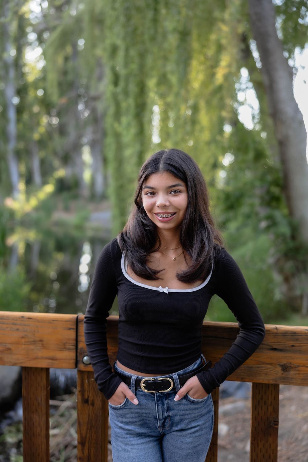Casual outdoor photoshoot on a wooden bench with a black long-sleeve top and jeans against a natural backdrop.
