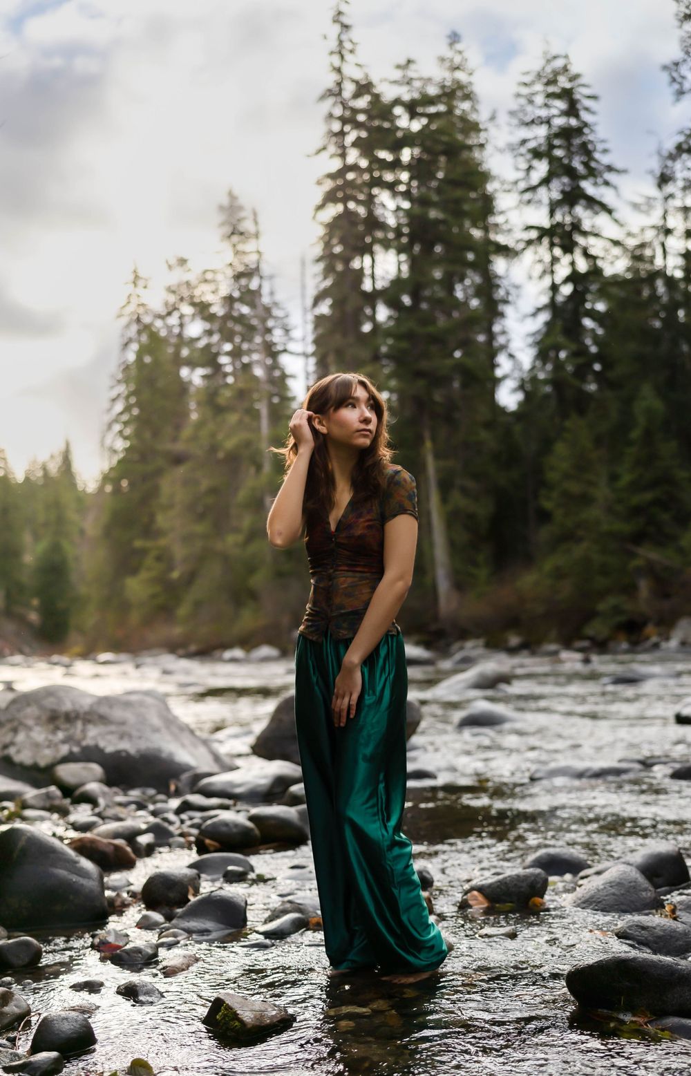 Person in emerald dress posing in flowing river surrounded by forest and rocks.