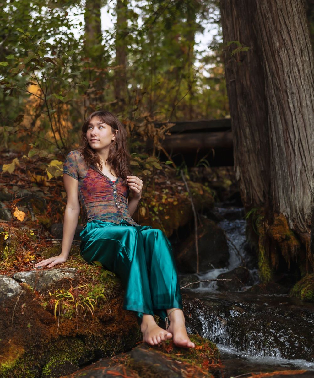 A whimsical forest photoshoot featuring a teal dress by a small waterfall among autumn leaves and mossy rocks.