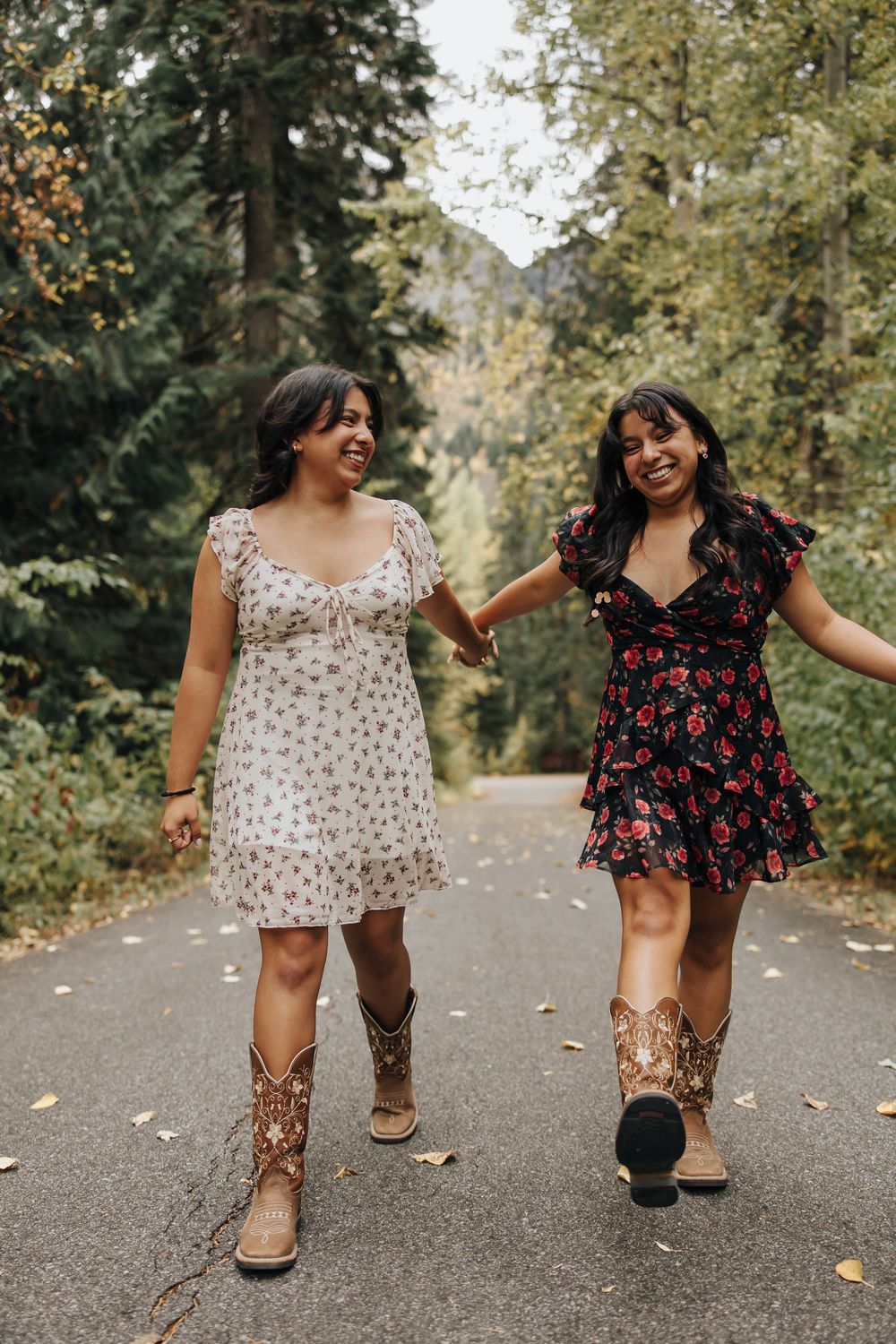 Two people in summer dresses walk together on a leaf-scattered path through the woods.
