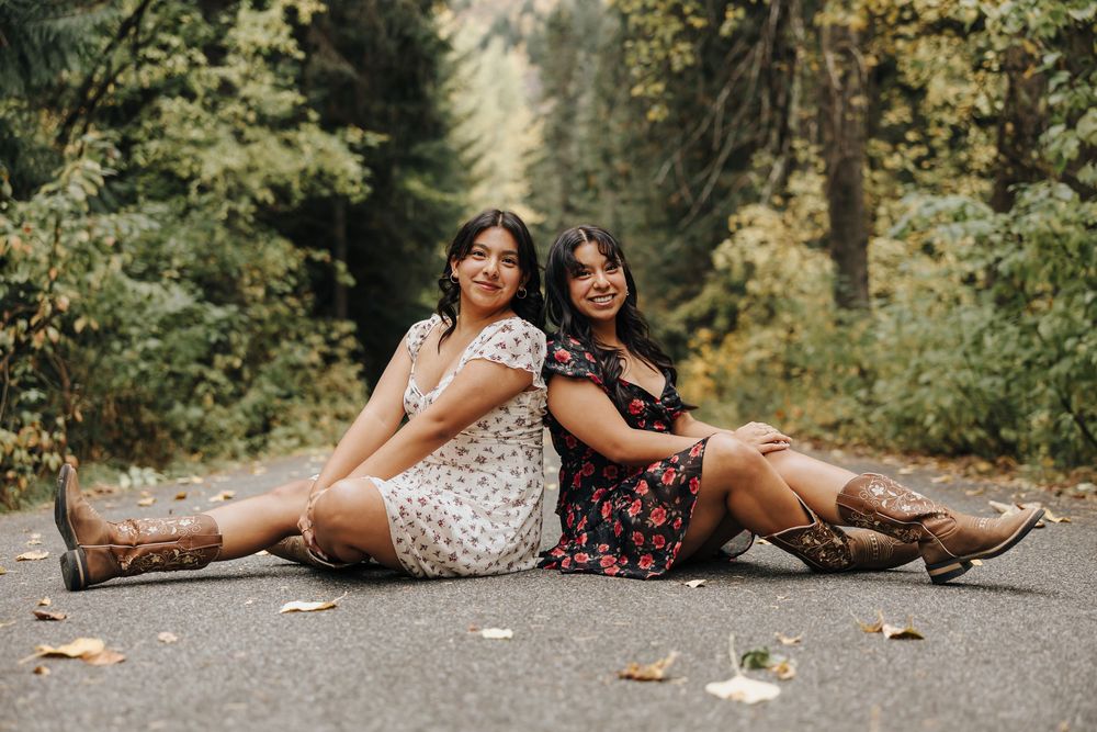 Two friends in floral dresses pose playfully on a forest path during autumn.