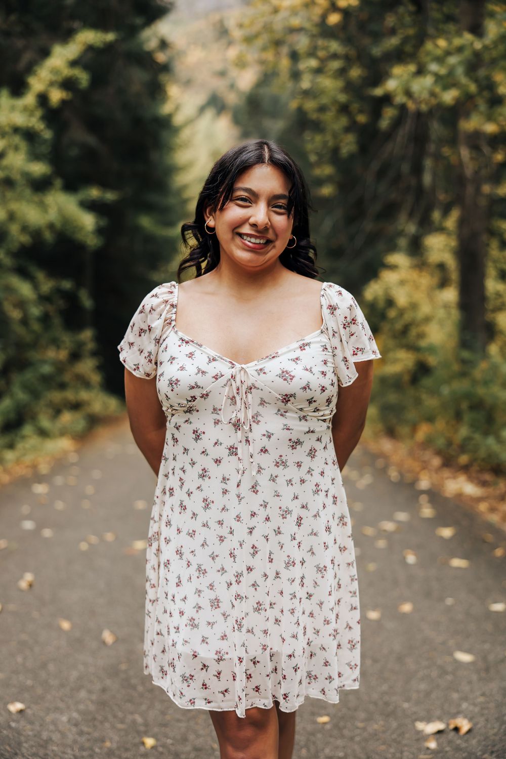 Person in a white floral dress poses on a wooded path during fall.