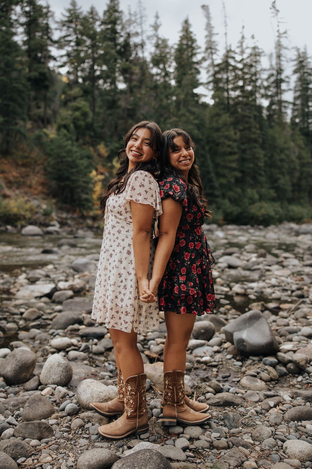 Two people in floral dresses pose together on rocky terrain with evergreen trees in background.