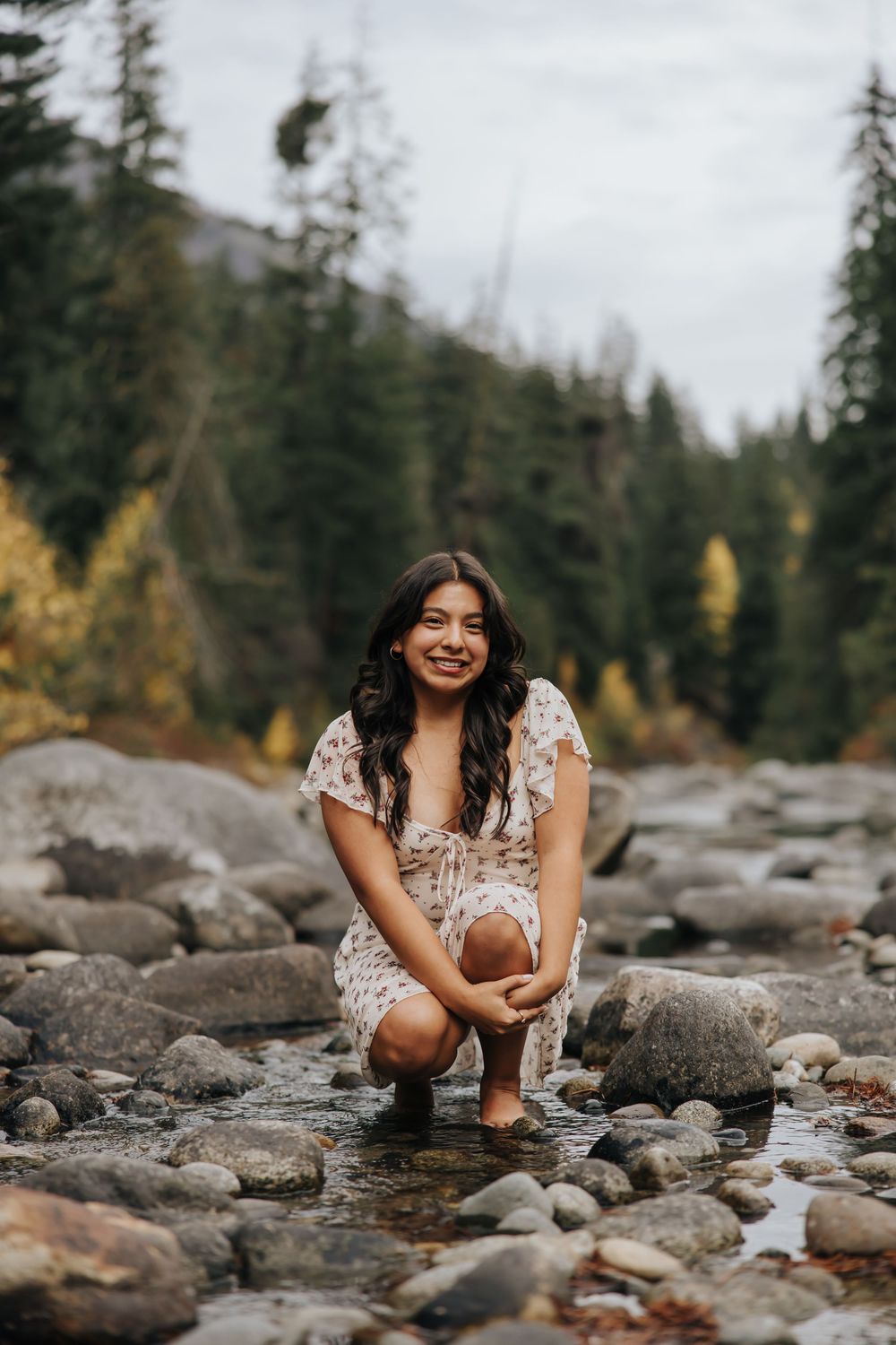 Person in white dress crouches by a mountain stream surrounded by evergreen forest.