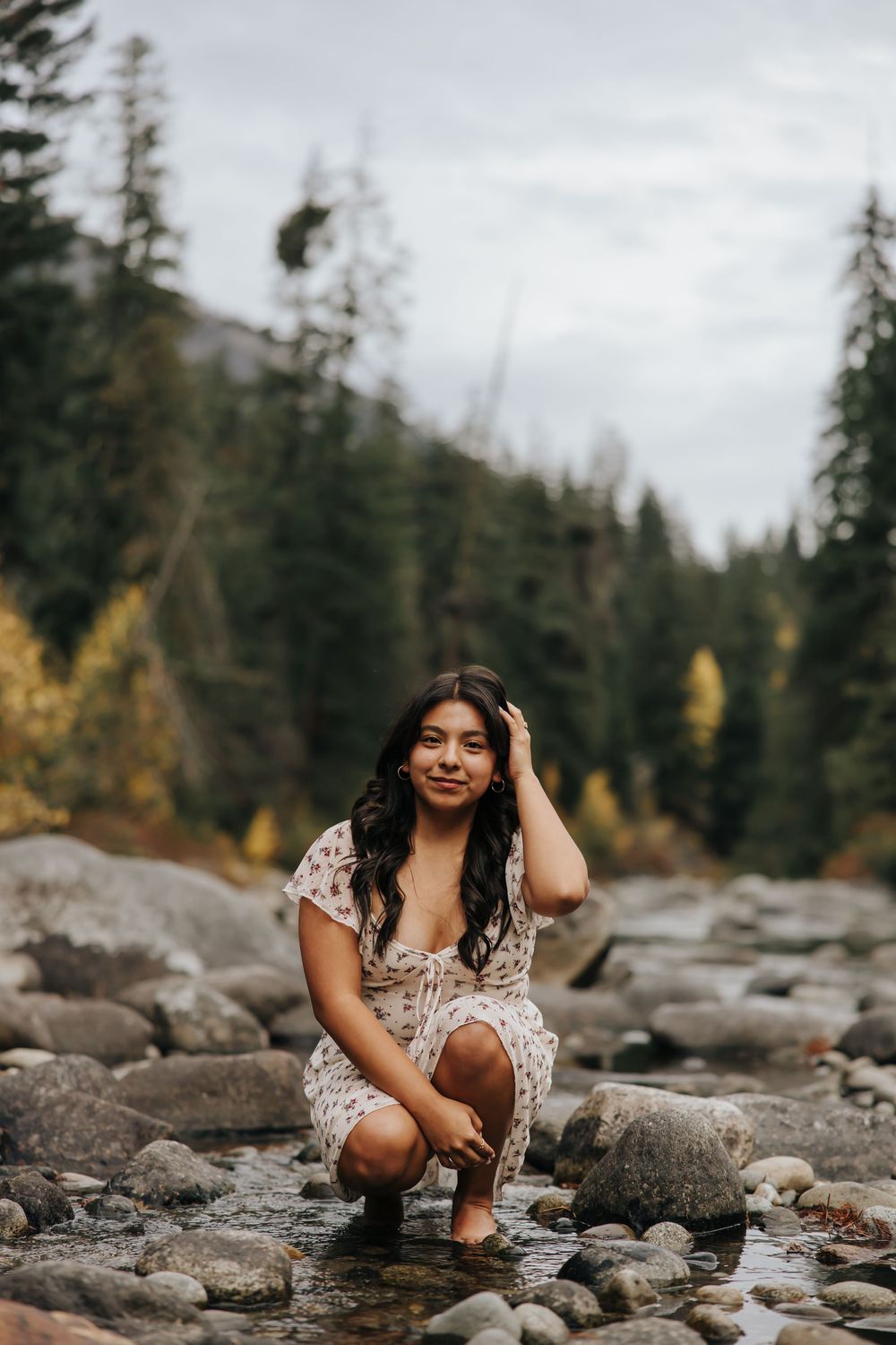 Person in floral dress poses among rocks in a scenic mountain stream setting.