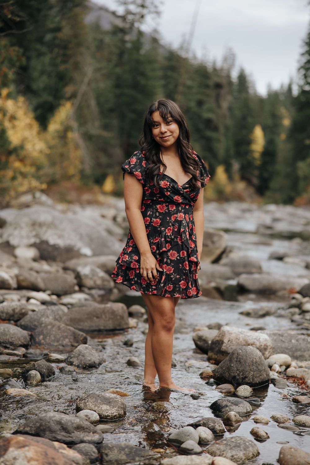 Person in red floral dress explores rocky mountain stream bed surrounded by evergreens.