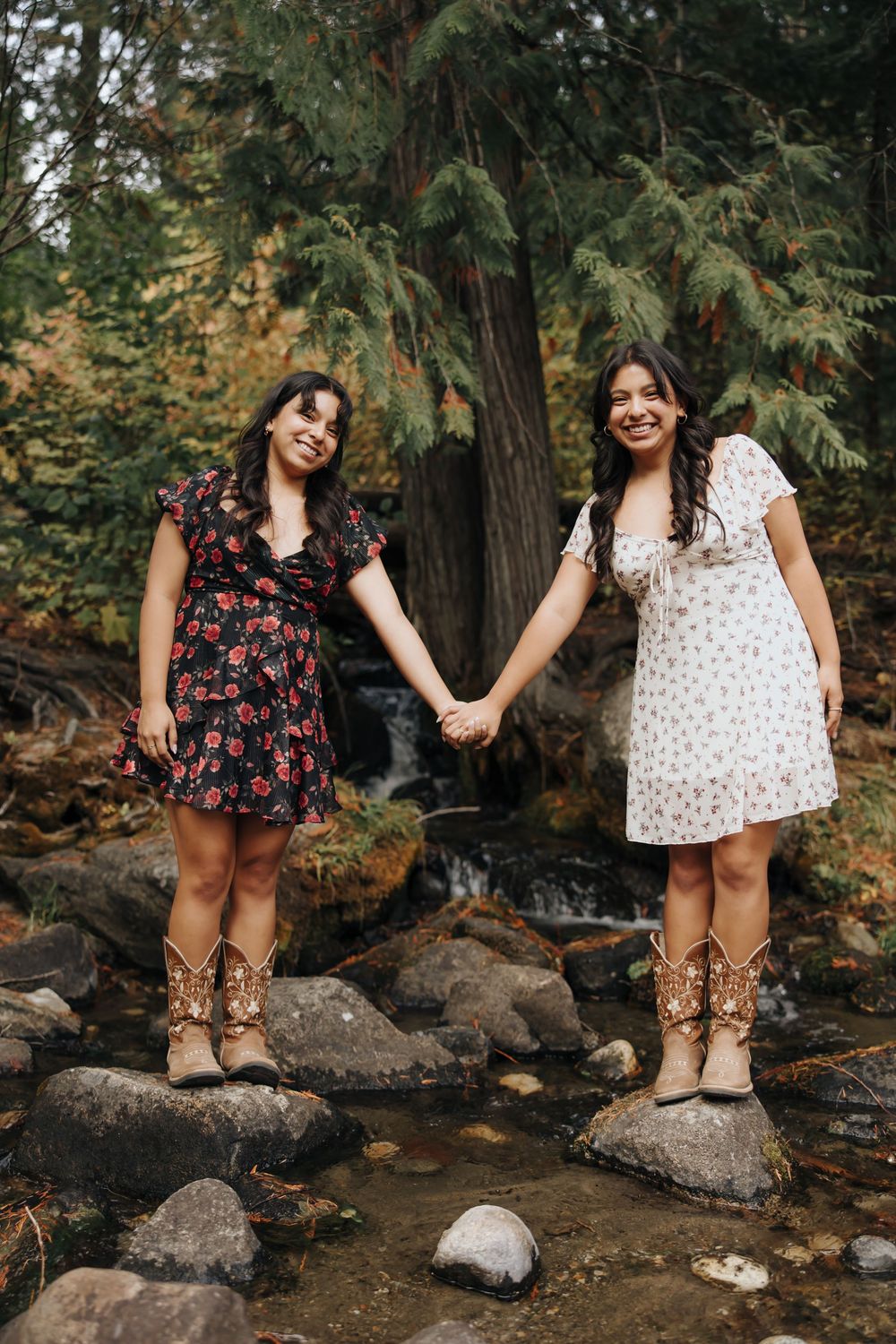 Two people in floral dresses hold hands while standing on rocks by a stream in a forest setting.
