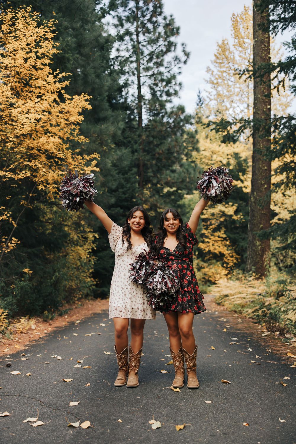 Two people in contrasting dresses hold pom poms high while posing on a fall forest path.