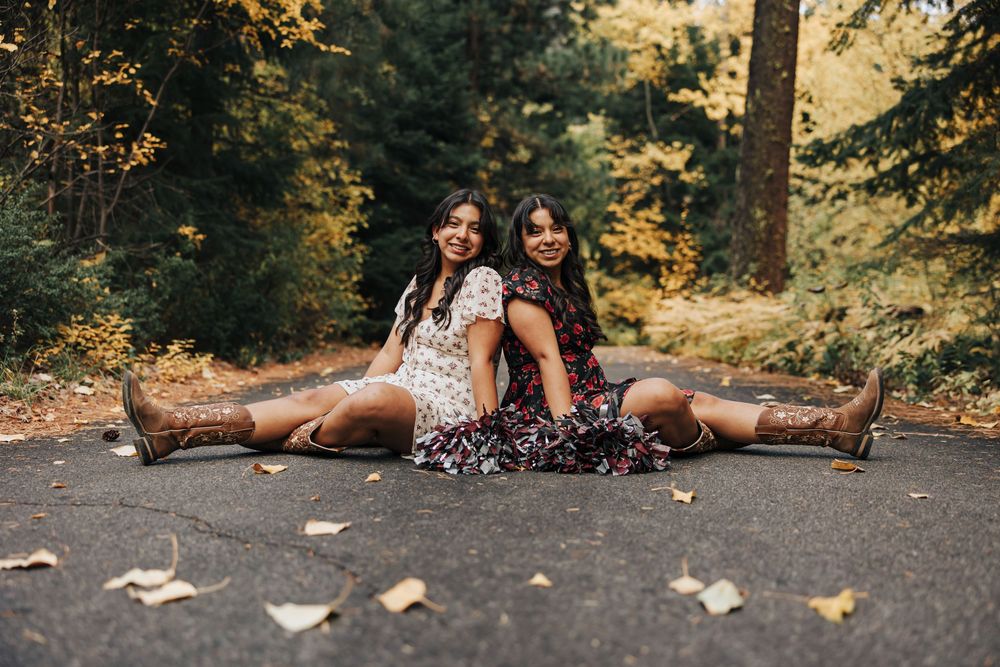 Two people in floral dresses sit back-to-back on a forest path covered in fallen leaves.