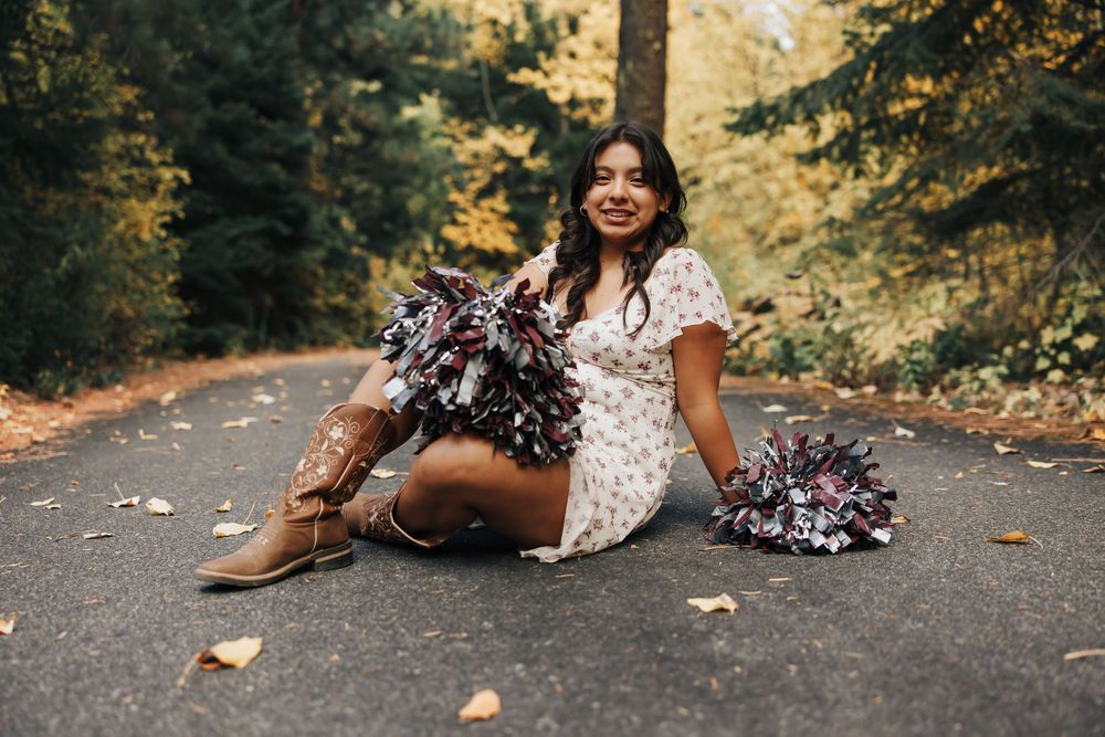 Person in white dress sits with pom poms on autumn forest path during photoshoot.