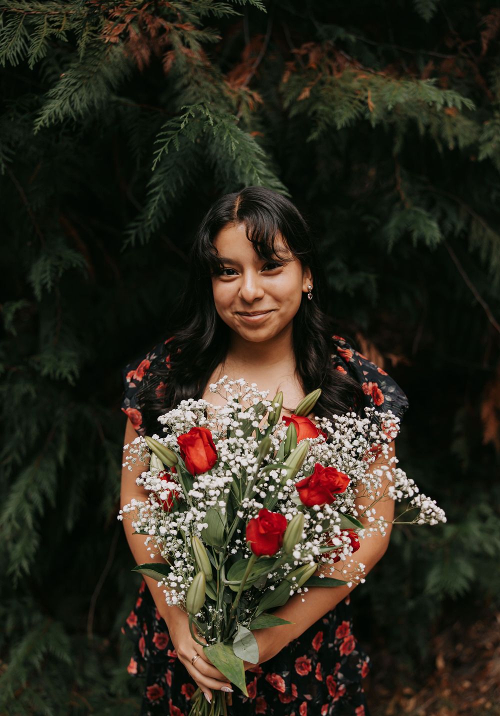 Person in floral dress holds bouquet of red roses and baby's breath against evergreen background.