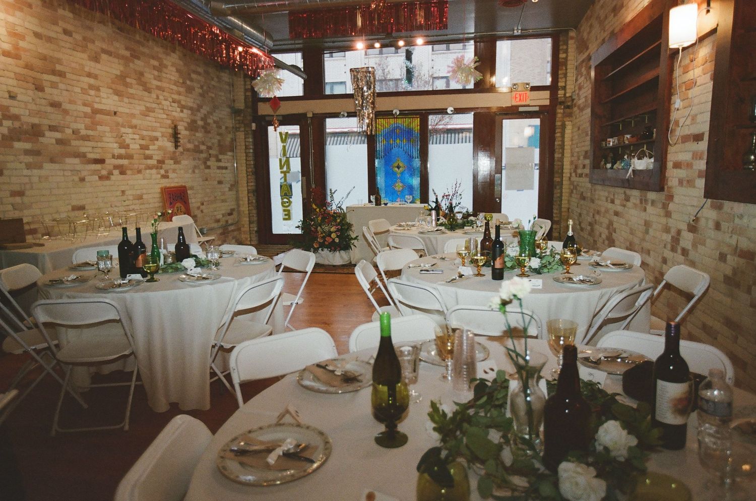 Rustic wedding reception venue with round tables, white linens, and exposed brick walls decorated with greenery and wine bottles.