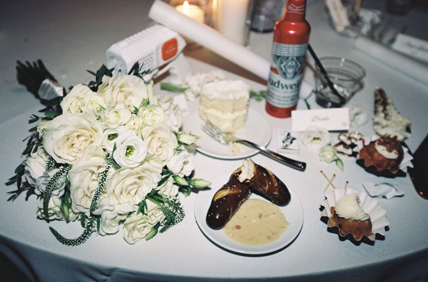 White rose bouquet on table with Budweiser beer, champagne glass, desserts, and candle at wedding reception.