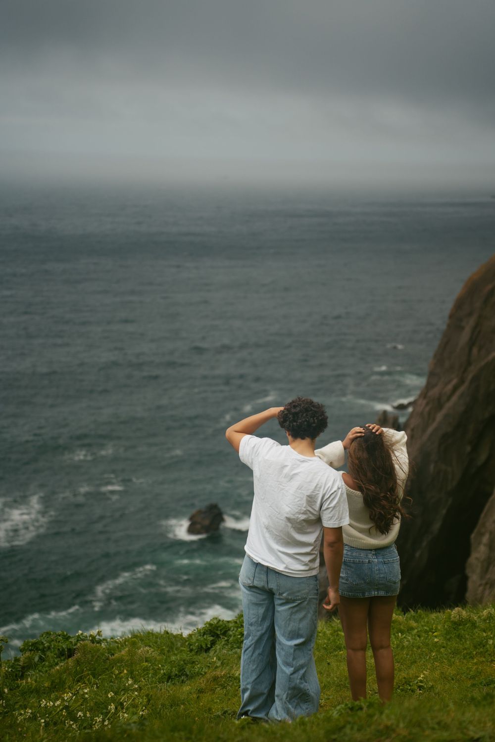 Couple standing on coastal cliff edge overlooking stormy ocean waves, viewed from behind on grassy overlook.