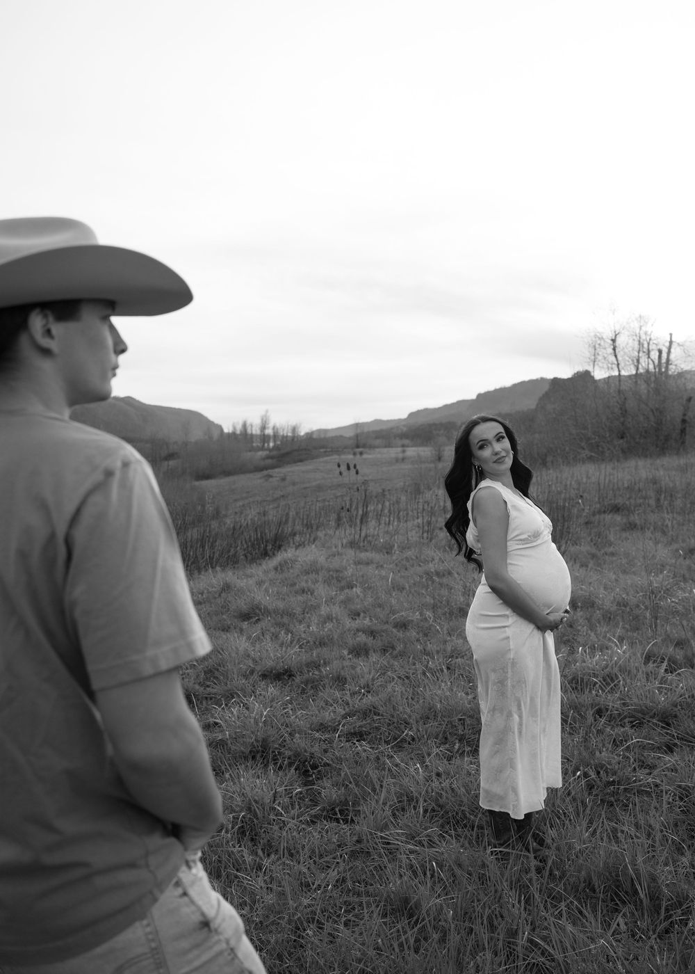Black and white maternity photo of pregnant woman in white dress smiling at man in cowboy hat in a rural field.