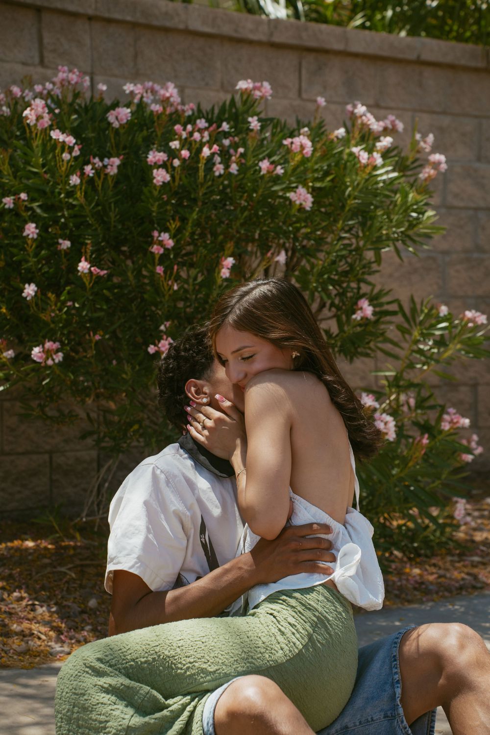Couple sharing a romantic embrace outdoors near blooming pink oleander bushes on a sunny day.