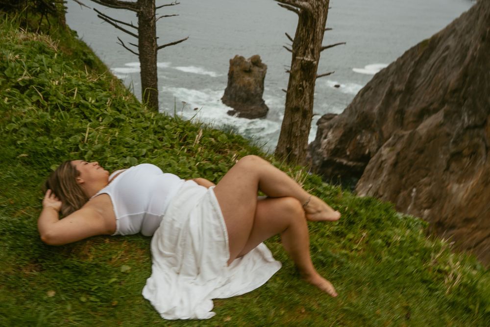 Pregnant woman in white dress lying on grassy coastal cliff with ocean rocks and waves in background.