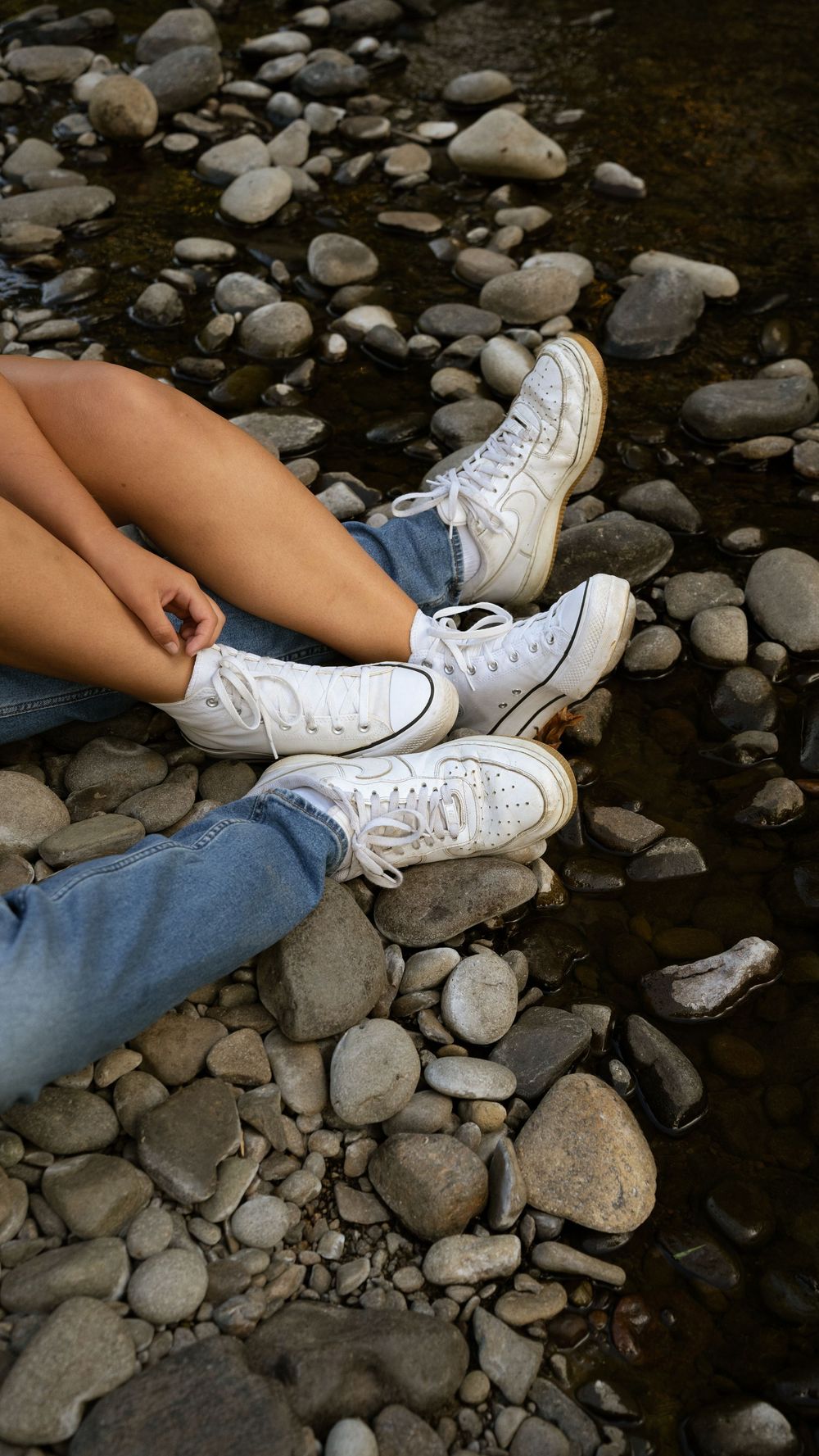 Person sitting on rocky riverbank wearing white high-top Converse sneakers and blue jeans.