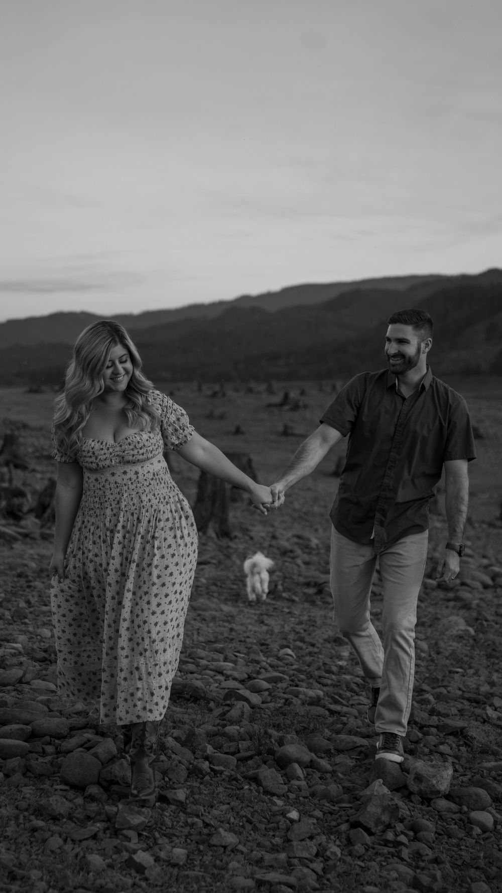 Black and white photo of a couple holding hands walking on rocky terrain with a dog and mountains in the background.