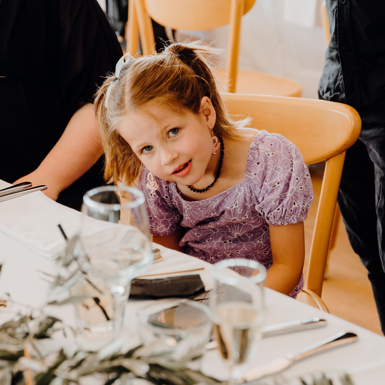 Child in sparkly pink dress sitting at dining table with place settings and glassware during a meal.