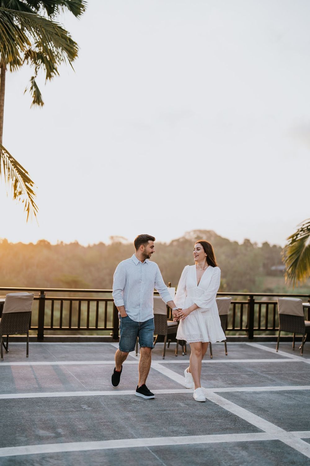 A couple walks together on a rooftop terrace during sunset with palm trees overhead.