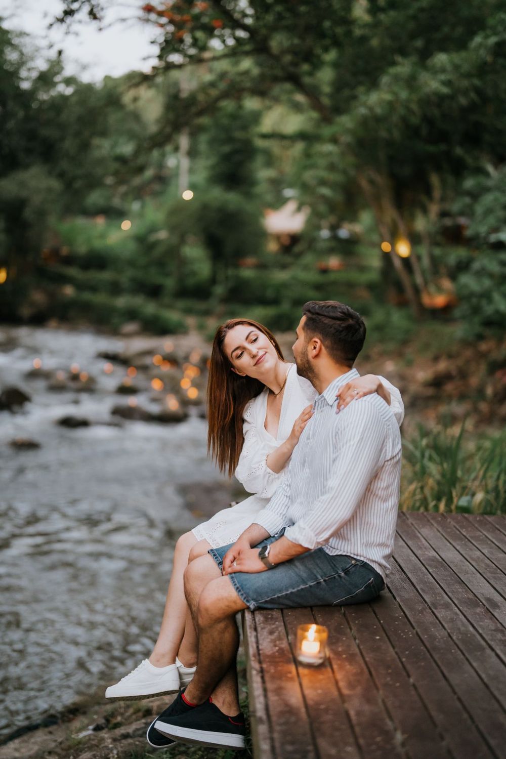 Intimate couple's portrait session on a wooden deck by a scenic river with glowing lanterns.