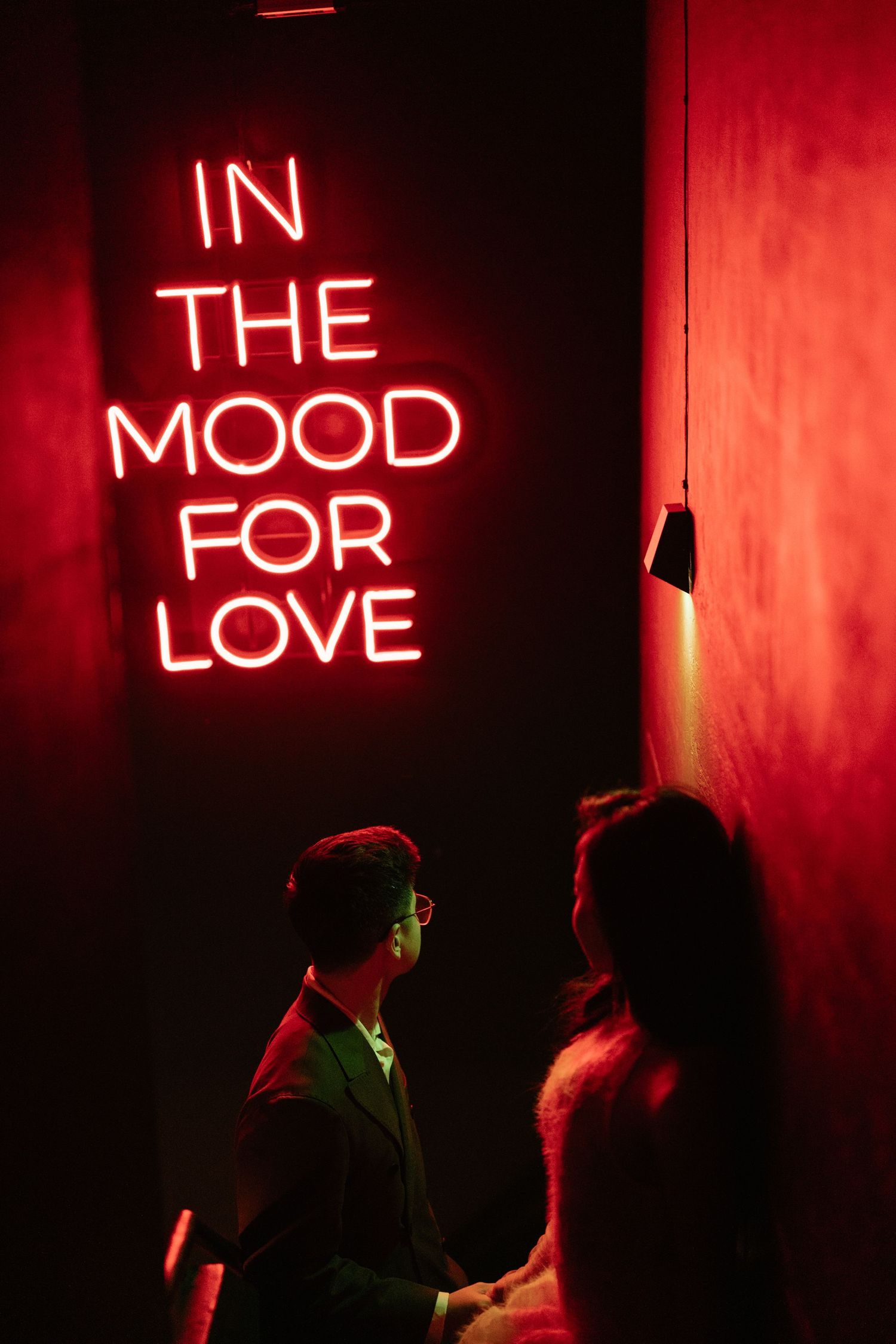 Couple standing near a glowing neon sign reading In The Mood For Love in a red-lit room.