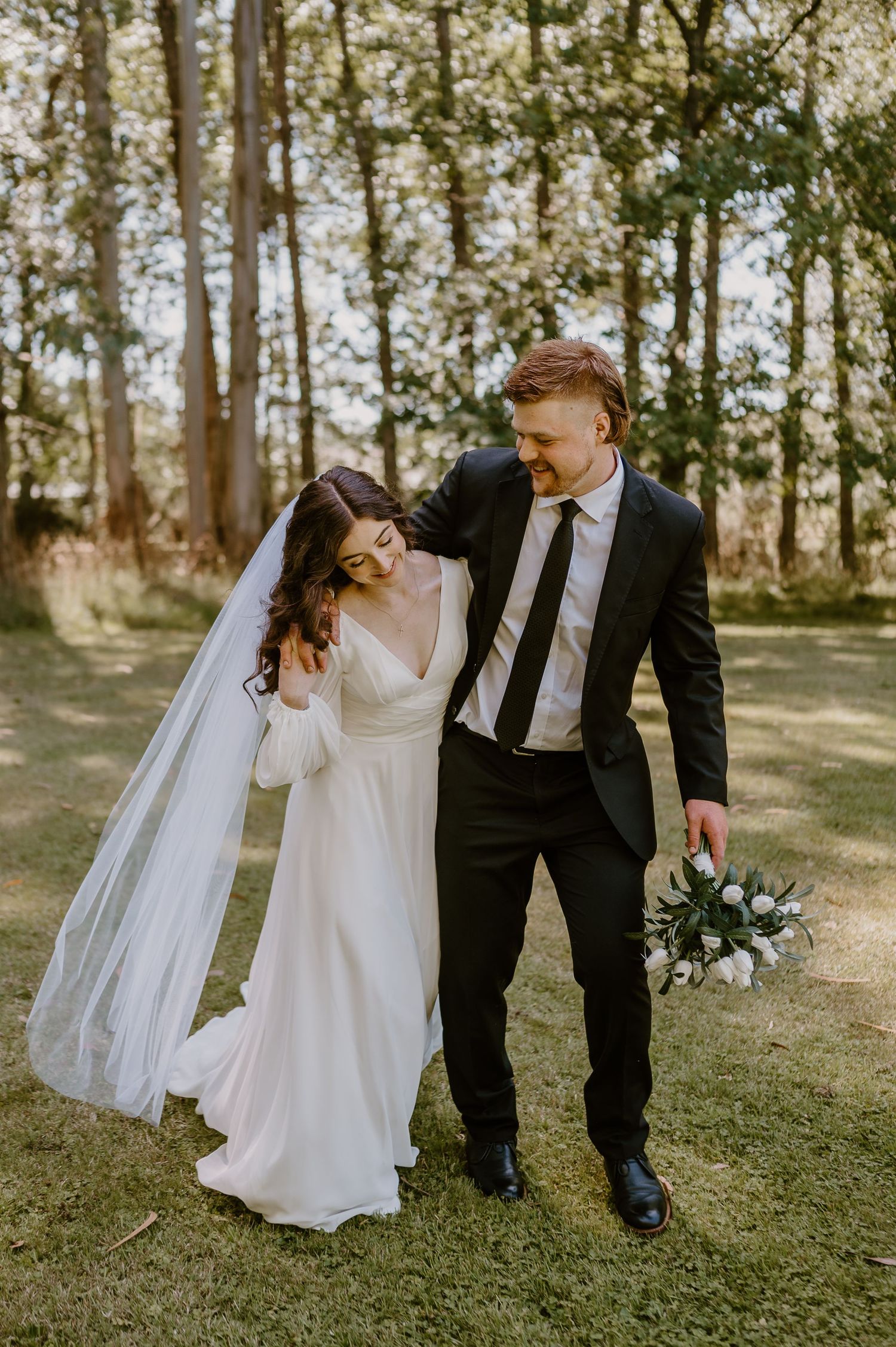 A bride and groom share an intimate moment while walking together in a sunlit wooded area in Christchurch.
