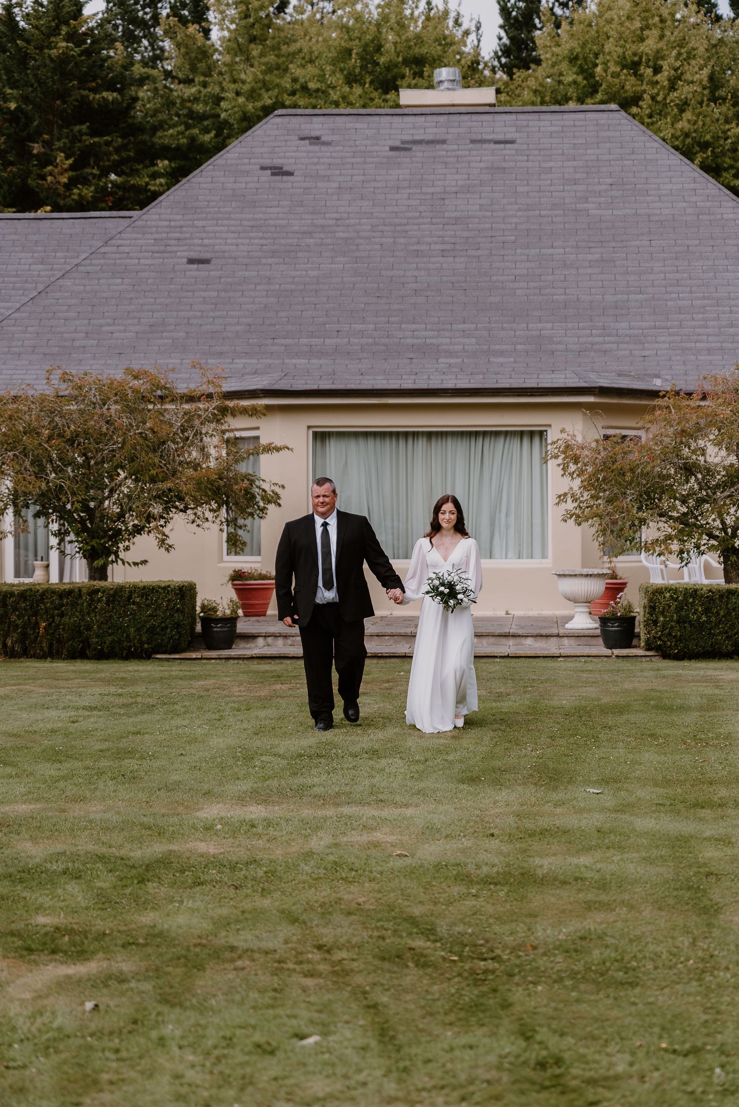 A couple walks together on a spacious green lawn in front of a ranch-style house during their wedding day. christchurch wedding photography