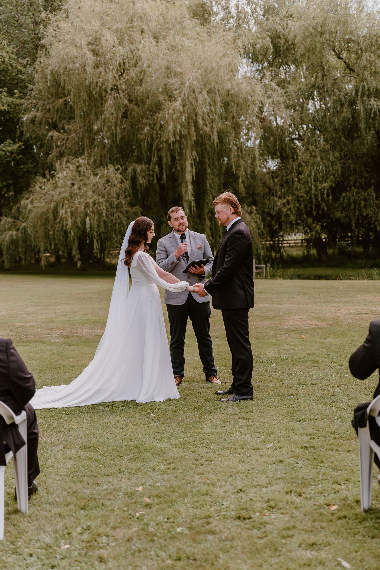 A wedding ceremony takes place outdoors in a field with willow trees creating a natural backdrop for the intimate gathering. christchurch
