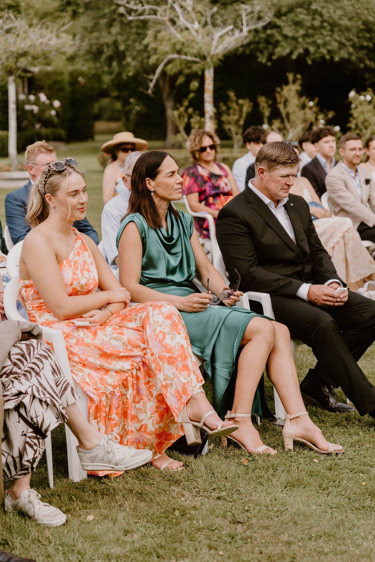 Wedding guests sit casually on white chairs on a lawn, with some wearing colorful summer dresses and formal attire. christchurch wedding