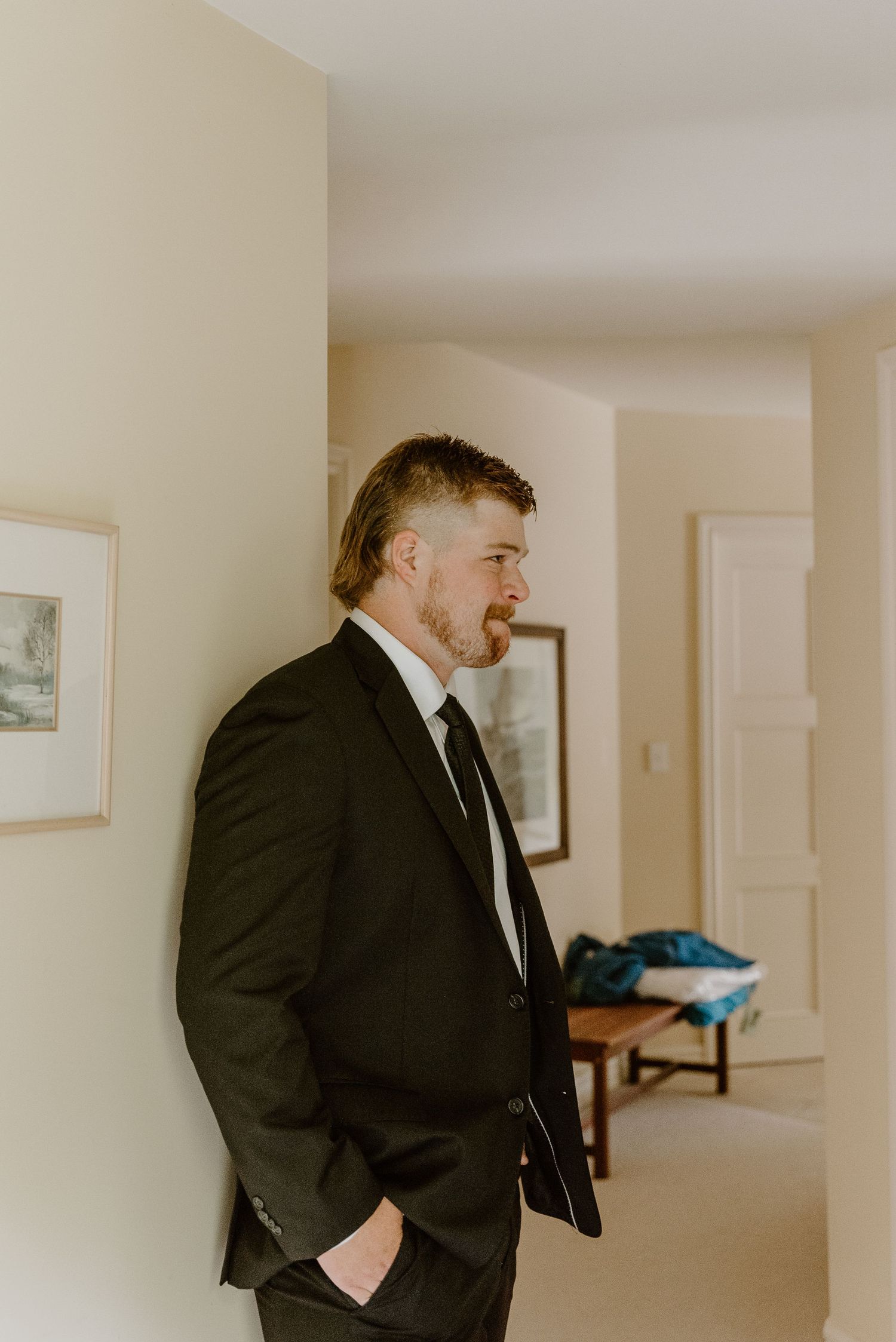 Man in black suit adjusts his tie while getting ready in a bright hallway with framed artwork. christchurch wedding photography