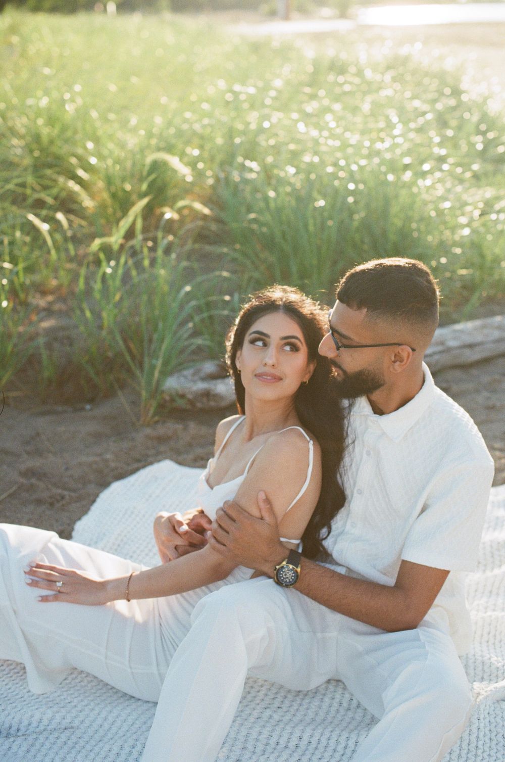 A couple in white outfits sharing romantic moments on a picnic blanket surrounded by sunlit grass.